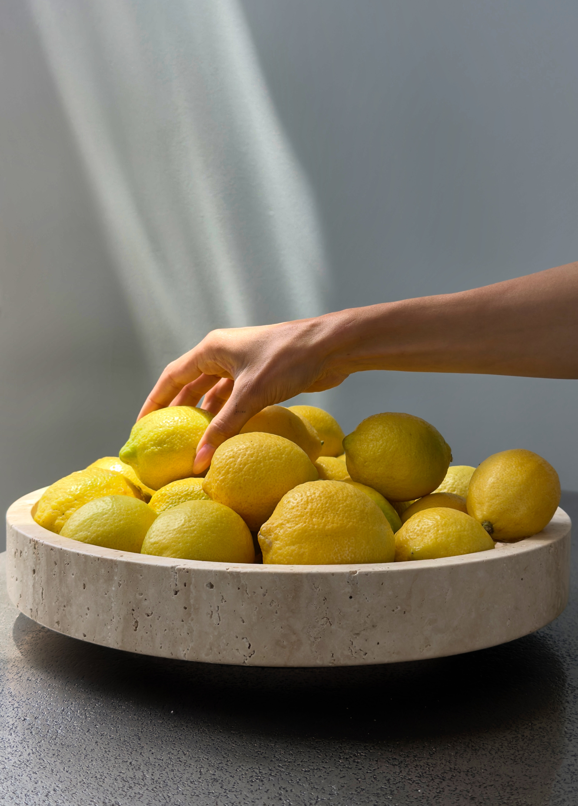 A hand reaching into a travertine bowl filled with whole lemons on a dark stone surface.